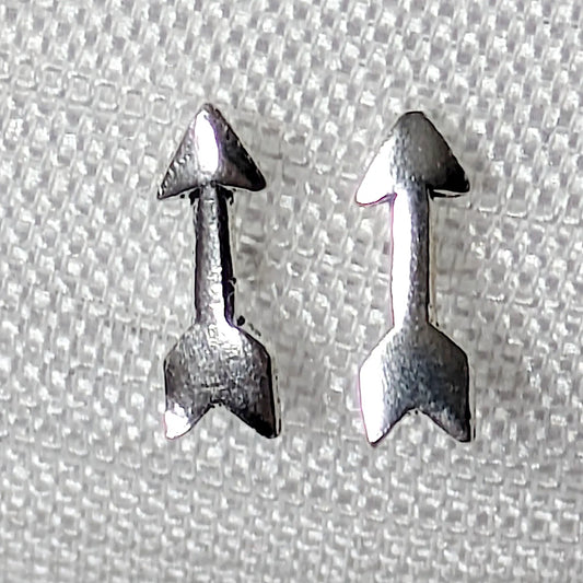 Nahaufnahme von zwei kleinen Pfeil-Ohrsteckern aus glänzendem 925 Silber auf hellem Stoffhintergrund.