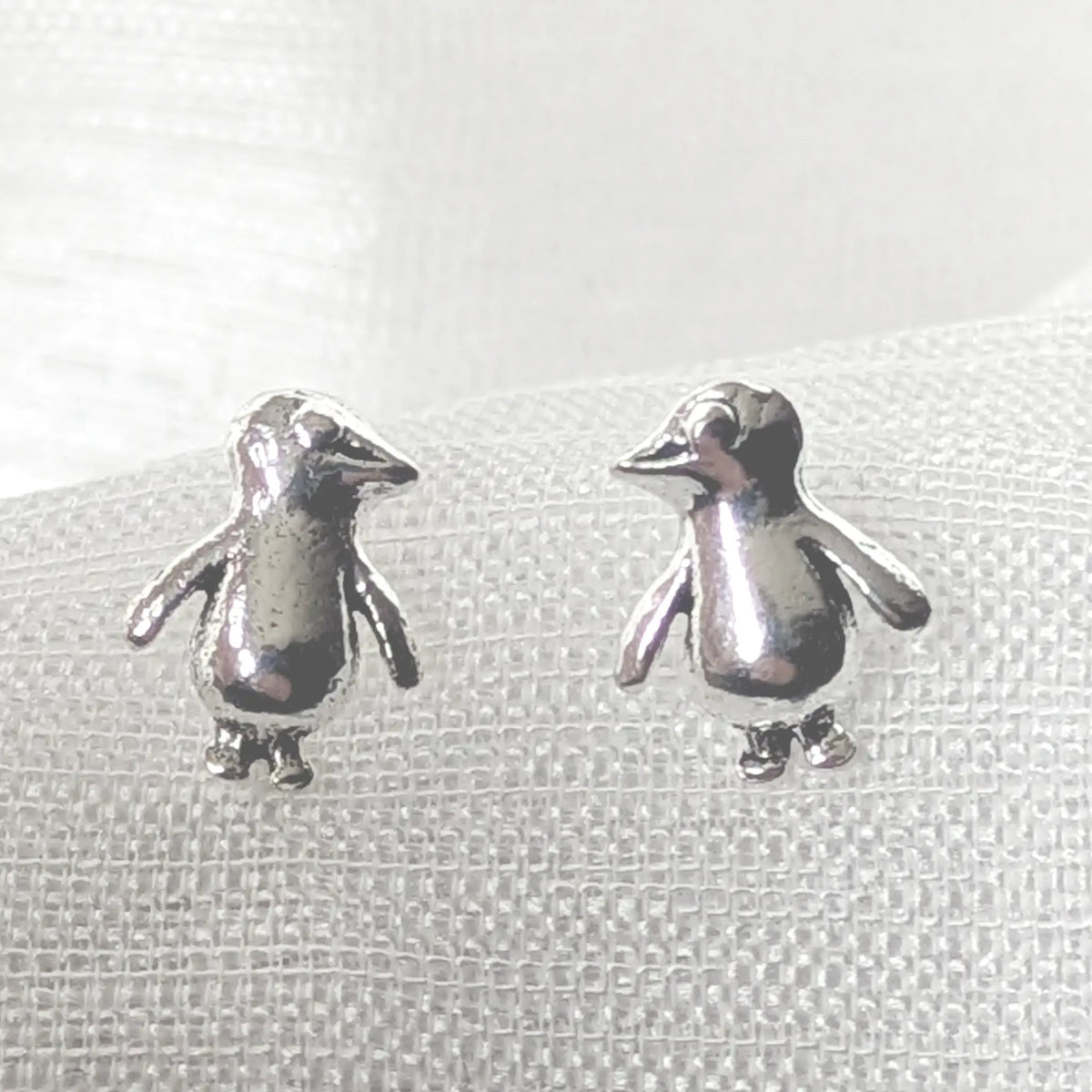 Detaillierte Nahaufnahme von zwei glänzenden 925 Silberohrsteckern in Form kleiner Pinguine, liegend auf einem hellen, strukturierten Stoff.
