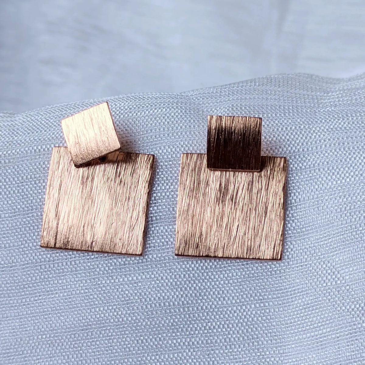 Nahaufnahme der geometrischen, rose-vergoldeten Ohrstecker aus 925 Silber mit zwei Quadraten und markant gebürsteter Struktur.