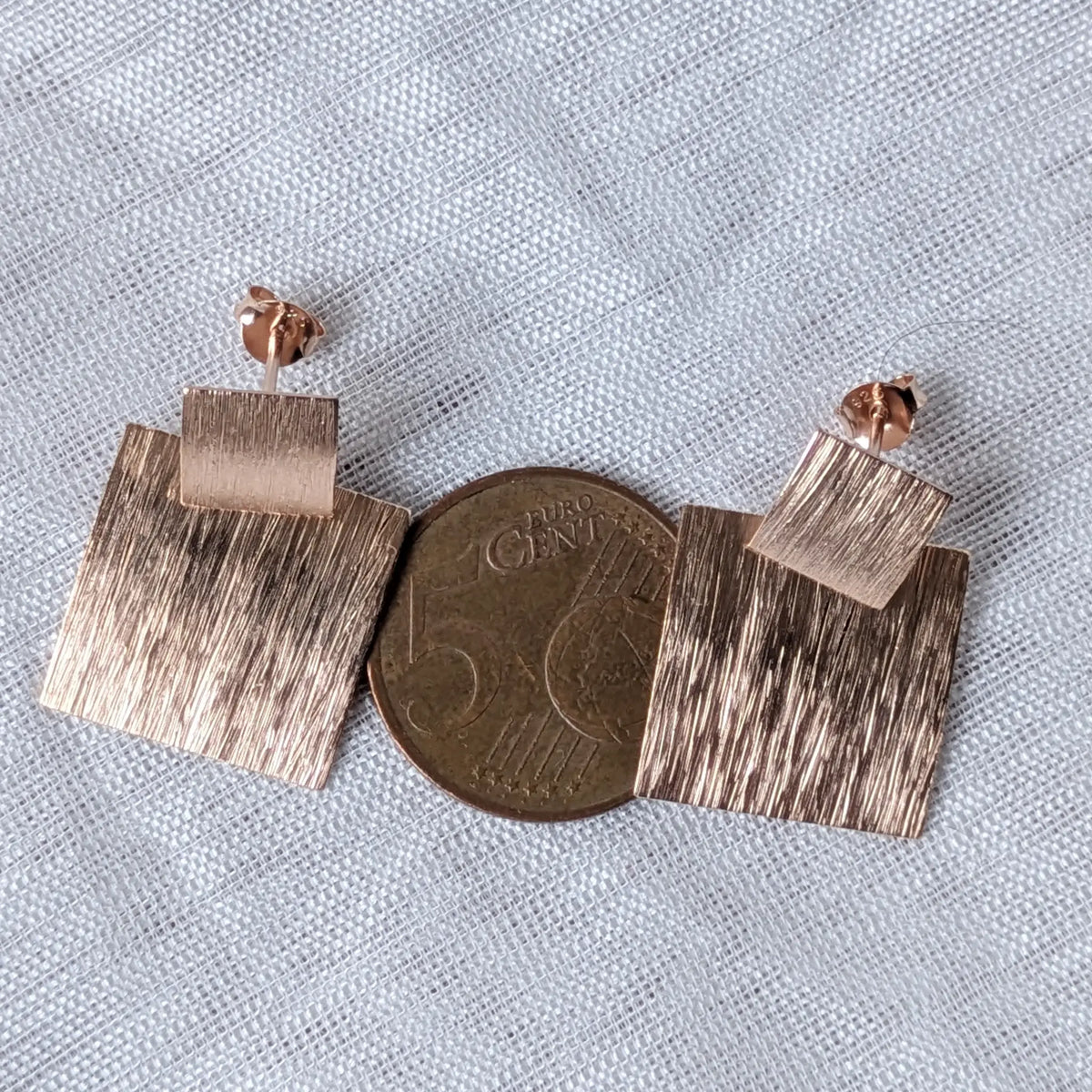 Größenvergleich der rose-vergoldeten eckigen Statement-Ohrringe, liegend auf einer 5-Cent-Münze zur Veranschaulichung der Dimensionen.
