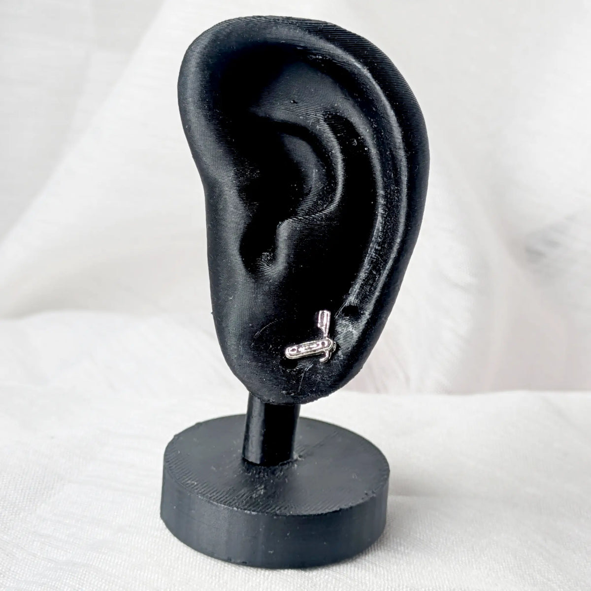 Ein silberner Rasiermesser-Ohrstecker, dezent getragen am Ohrläppchen eines schwarzen 3D-Modell-Ohrs.