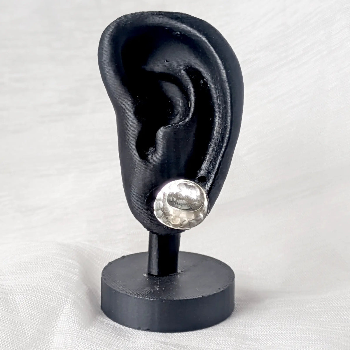 Ein runder, großer Silberohrstecker an einem schwarzen Ohr-Display zur Veranschaulichung der auffälligen Größe am Ohrläppchen.