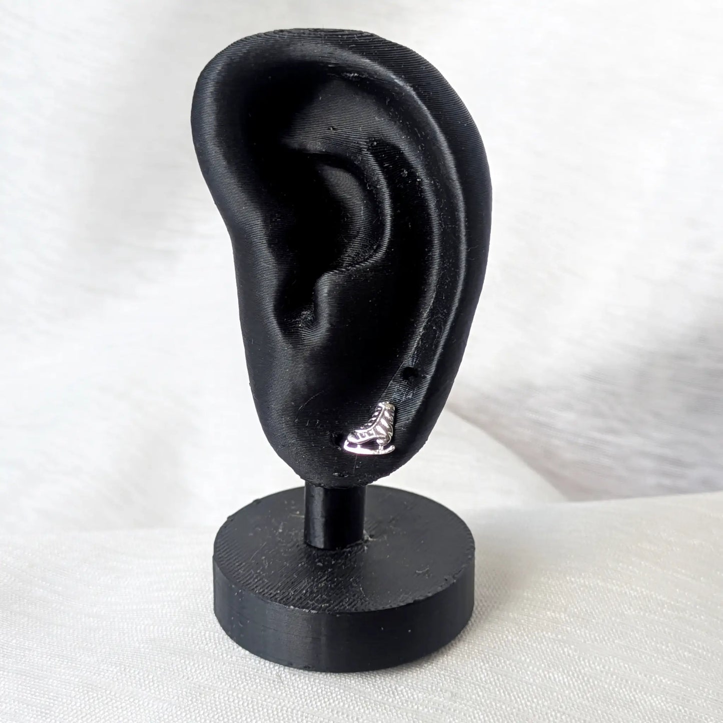 Ein filigraner Schlittschuh-Ohrstecker aus Silber getragen an einem mattschwarzen Deko-Ohr zur Veranschaulichung der Größe.