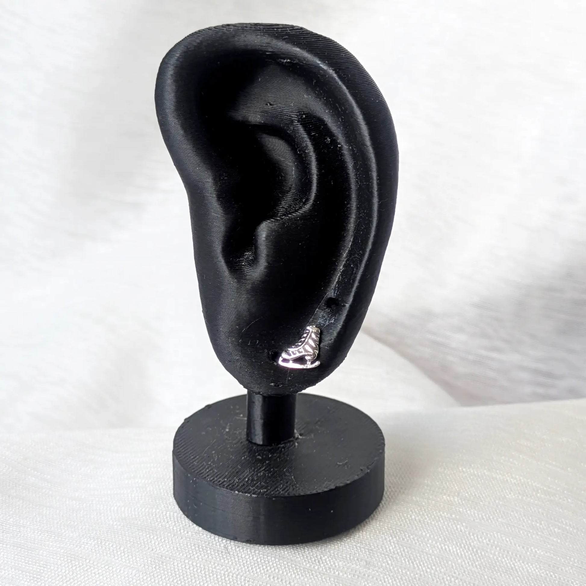 Ein filigraner Schlittschuh-Ohrstecker aus Silber getragen an einem mattschwarzen Deko-Ohr zur Veranschaulichung der Größe.