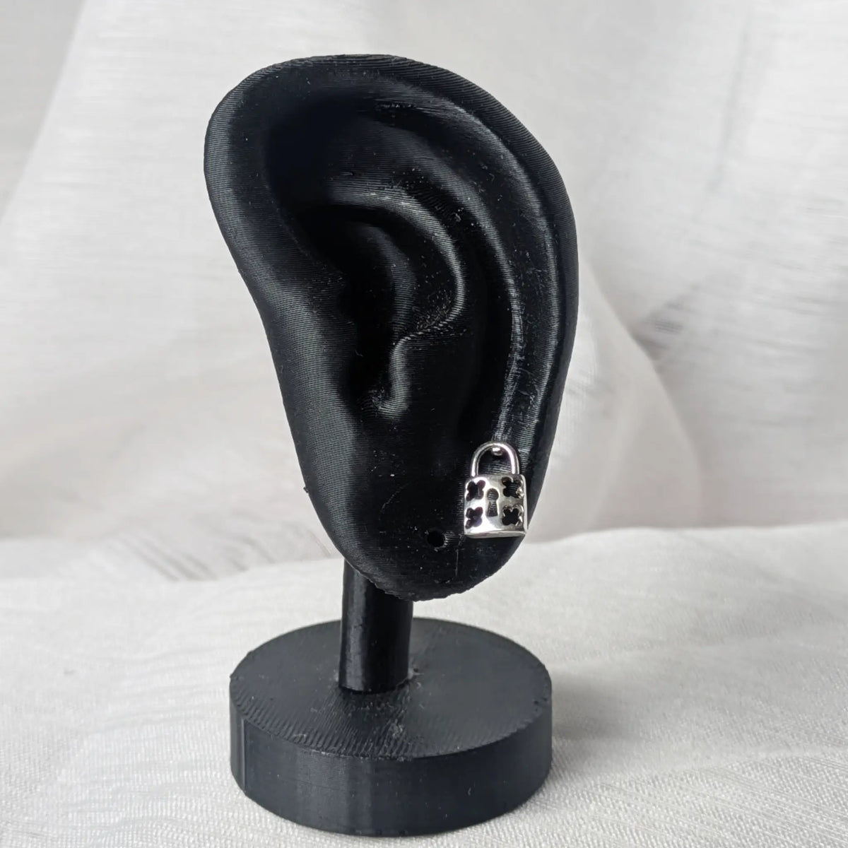 Der silberne Vorhängeschloss-Ohrstecker, dezent getragen am Ohrläppchen eines schwarzen 3D-Modell-Ohrs zur Größenveranschaulichung.
