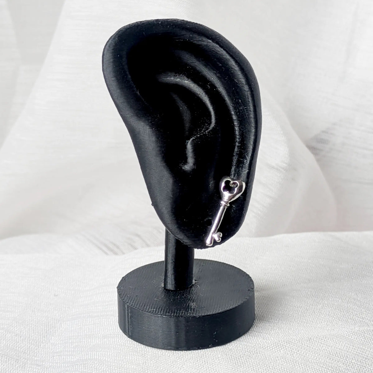 Der silberne Schlüssel-Ohrstecker, dezent getragen am Ohrläppchen eines schwarzen 3D-Modell-Ohrs zur Größenveranschaulichung.

