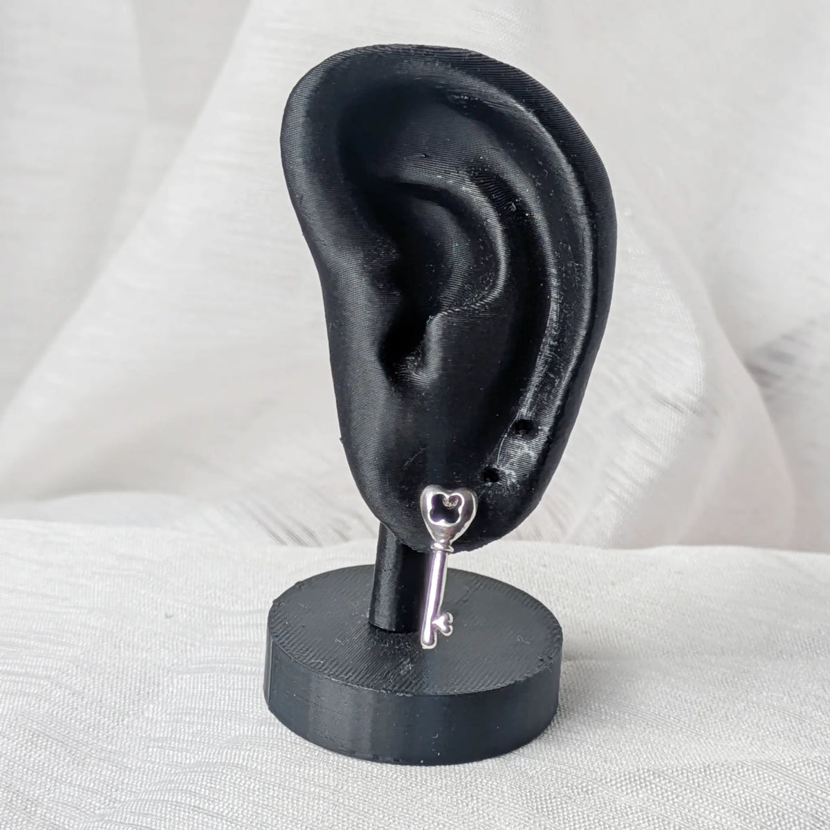 Der silberne Schlüssel-Ohrstecker, dezent getragen am Ohrläppchen eines schwarzen 3D-Modell-Ohrs zur Größenveranschaulichung