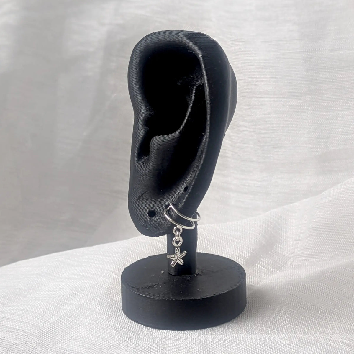 Eine silberne Seestern-Ohrklemme befestigt am unteren Ohrläppchen eines schwarzen 3D-Ohr-Displays zur Größendarstellung.