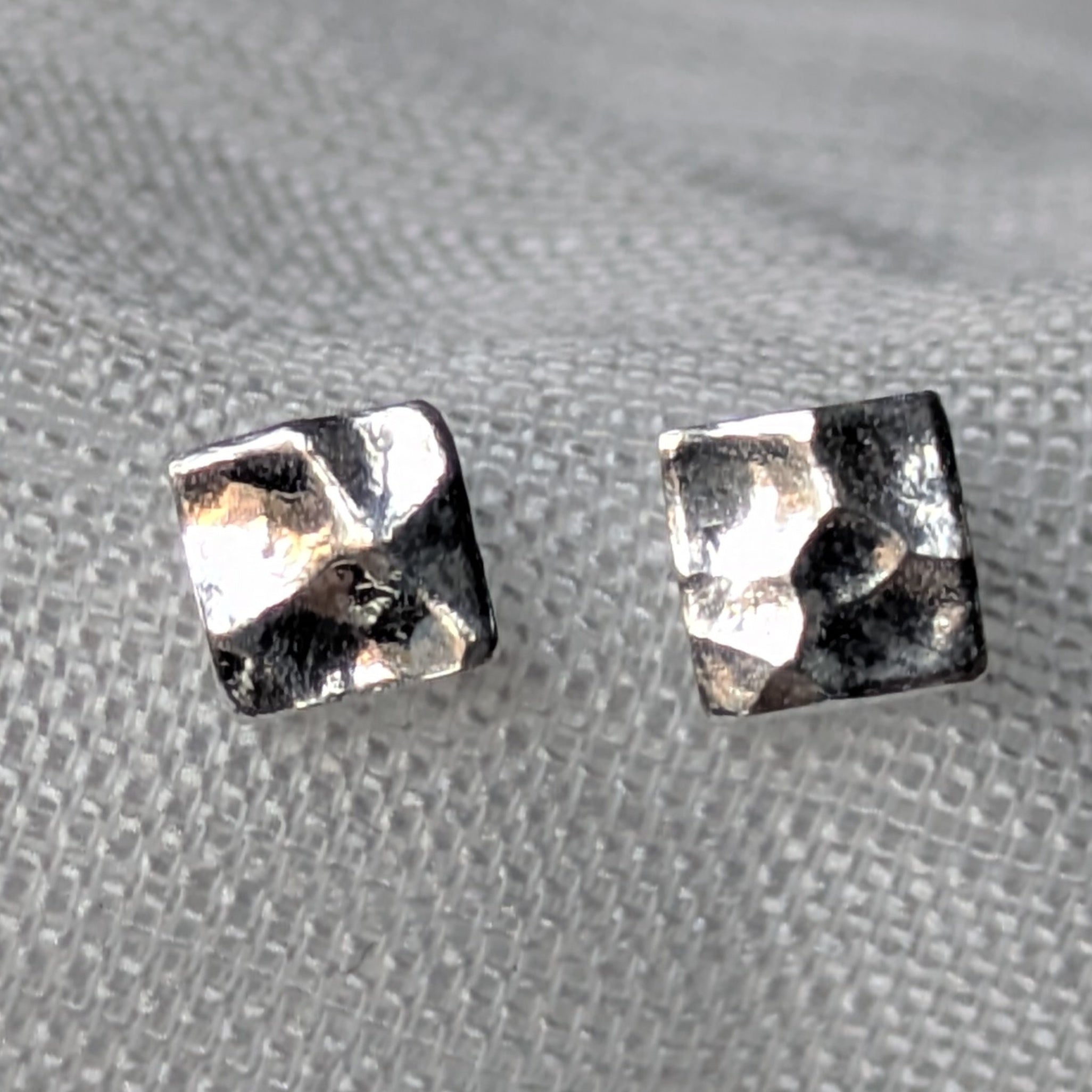 Quadratische, gehämmerte 925-Silber Ohrstecker auf heller Stoffoberfläche in Nahaufnahme.