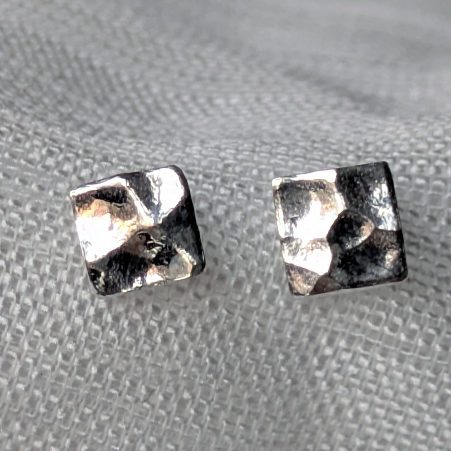 Quadratische, gehämmerte 925-Silber Ohrstecker auf heller Stoffoberfläche in Nahaufnahme.
