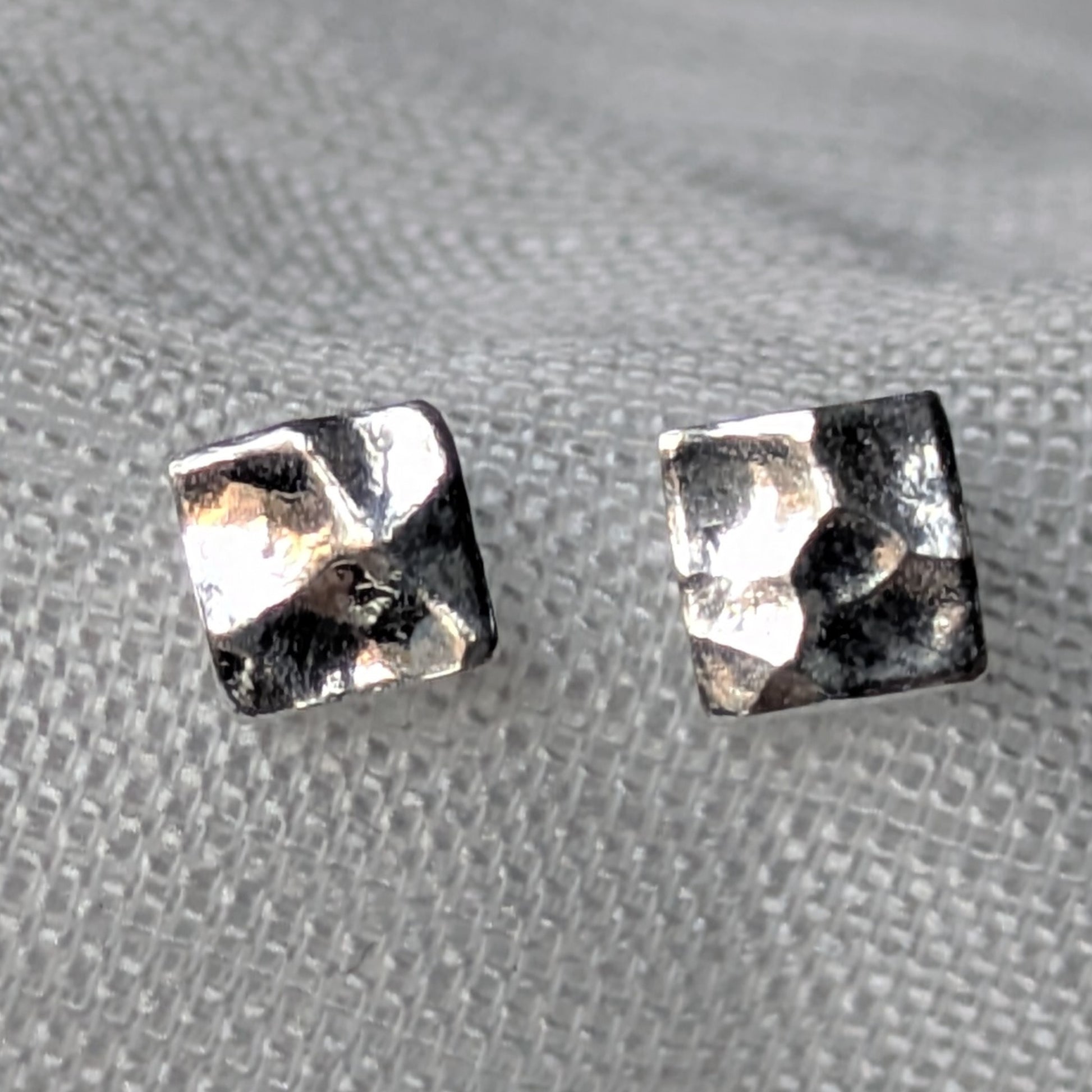 Quadratische, gehämmerte 925-Silber Ohrstecker auf heller Stoffoberfläche in Nahaufnahme.