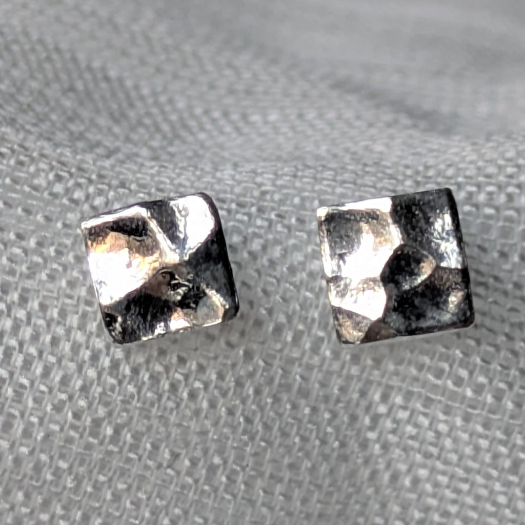 Quadratische, gehämmerte 925-Silber Ohrstecker auf heller Stoffoberfläche in Nahaufnahme.