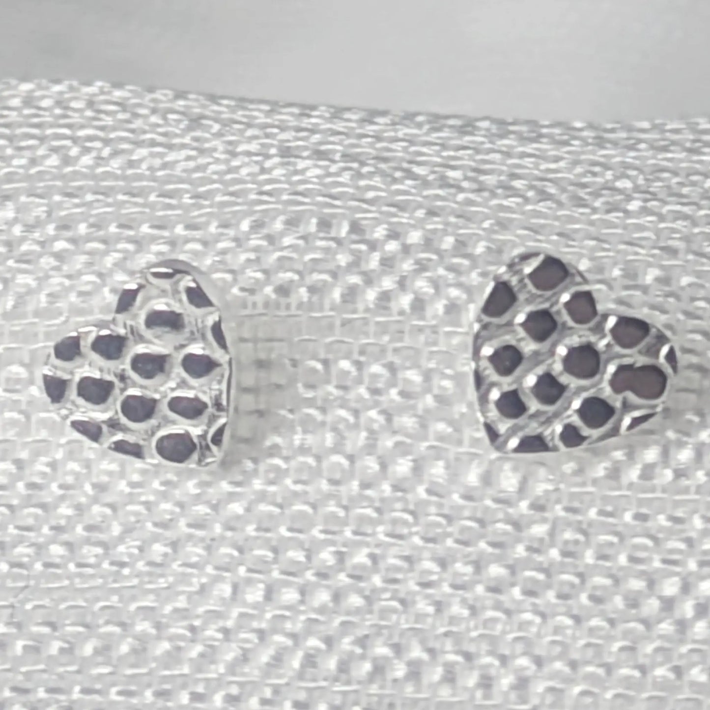 Detaillierte Nahaufnahme von zwei Herz-Ohrsteckern aus 925 Sterling Silber mit einer markanten, gehämmerten Wabenstruktur auf hellem Stoff.