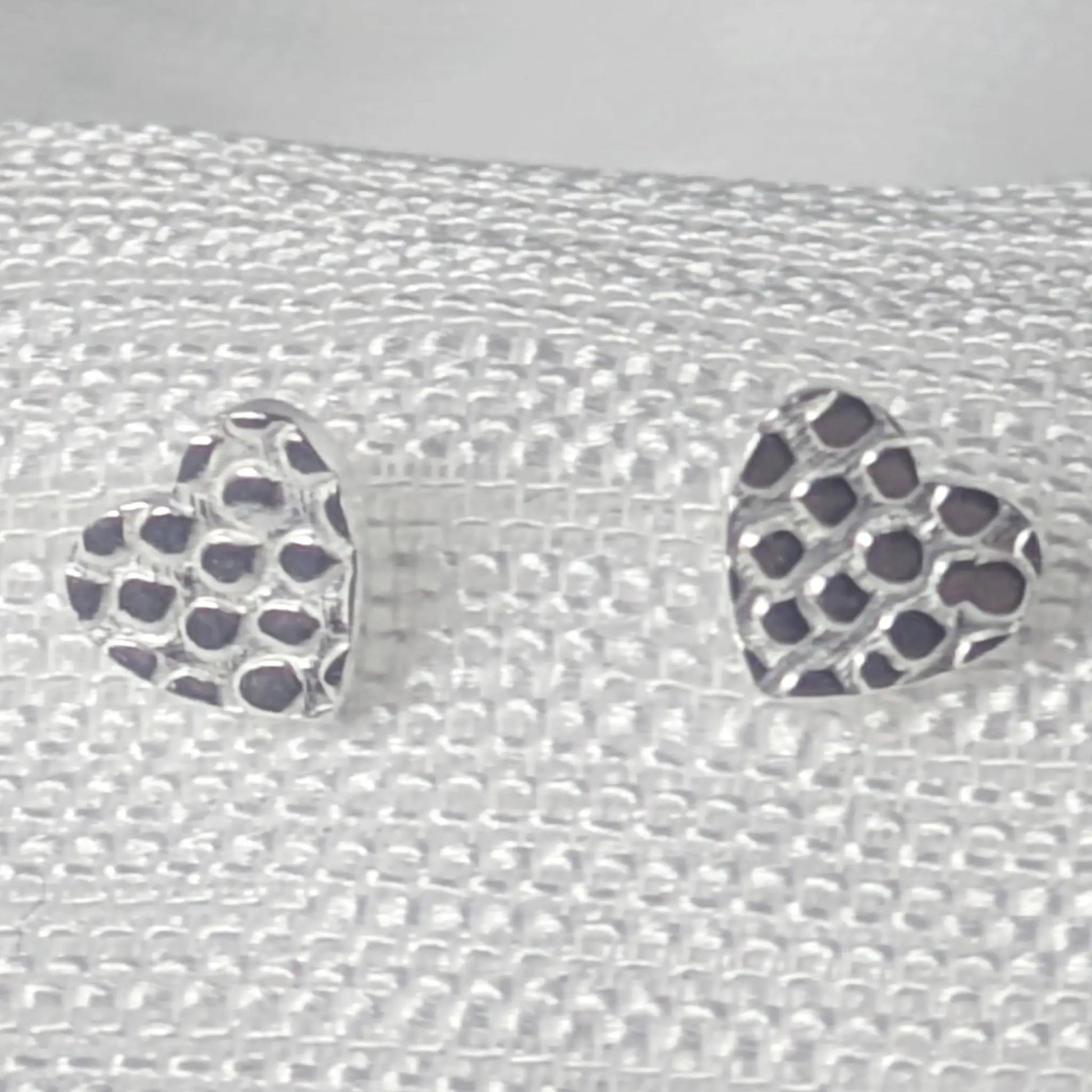 Detaillierte Nahaufnahme von zwei Herz-Ohrsteckern aus 925 Sterling Silber mit einer markanten, gehämmerten Wabenstruktur auf hellem Stoff.