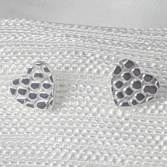 Detaillierte Nahaufnahme von zwei Herz-Ohrsteckern aus 925 Sterling Silber mit einer markanten, gehämmerten Wabenstruktur auf hellem Stoff.