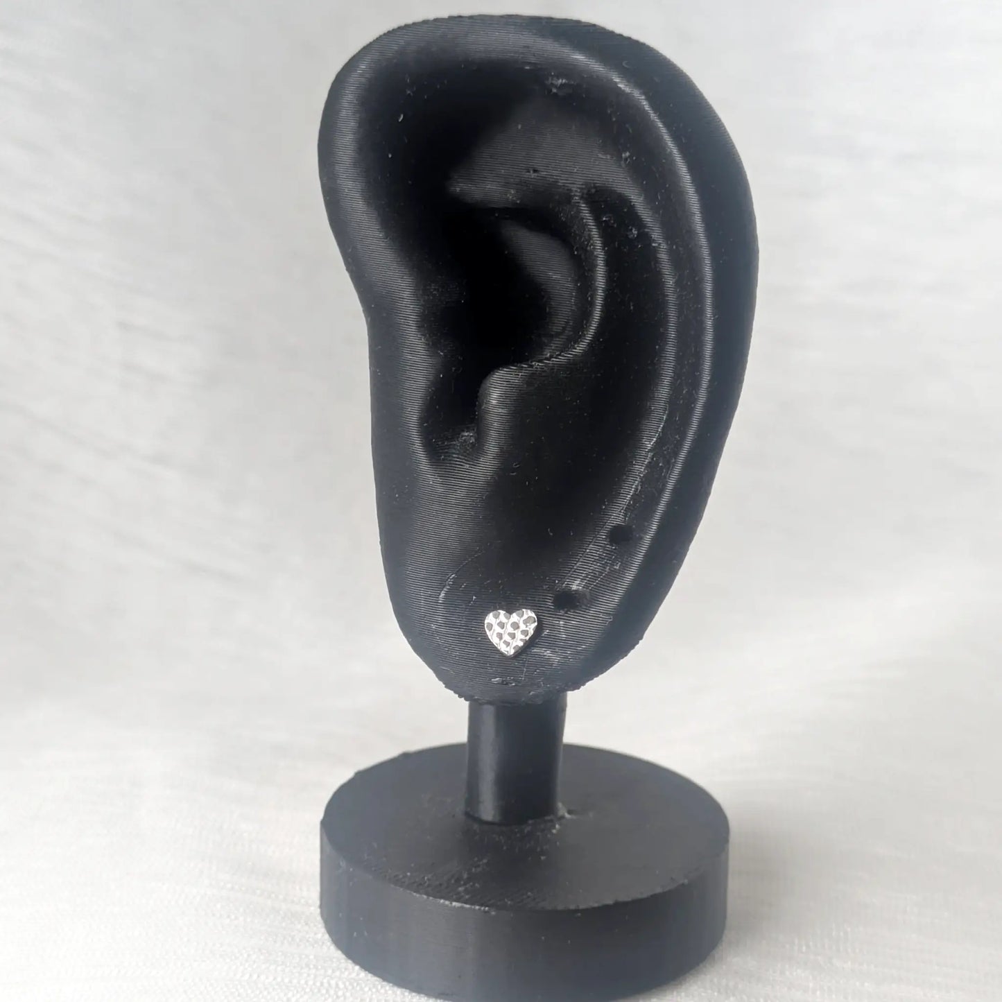 Ein einzelner gehammerter Herz-Ohrstecker aus Silber präsentiert an einem schwarzen Ohr-Modell zeigt die zarte, moderne Wirkung beim Tragen.