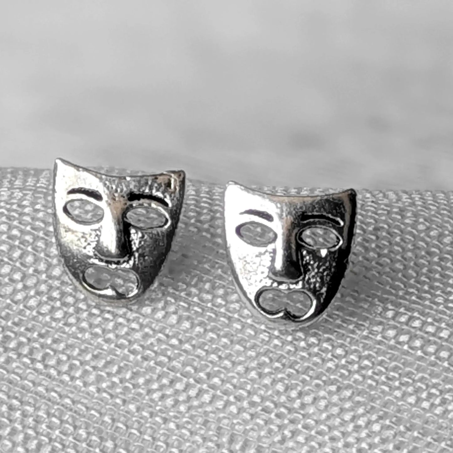 Detailreiche Nahaufnahme von zwei hochglanzpolierten Theatermasken Ohrsteckern aus 925 Sterling Silber mit dem Motiv der Tragödie.