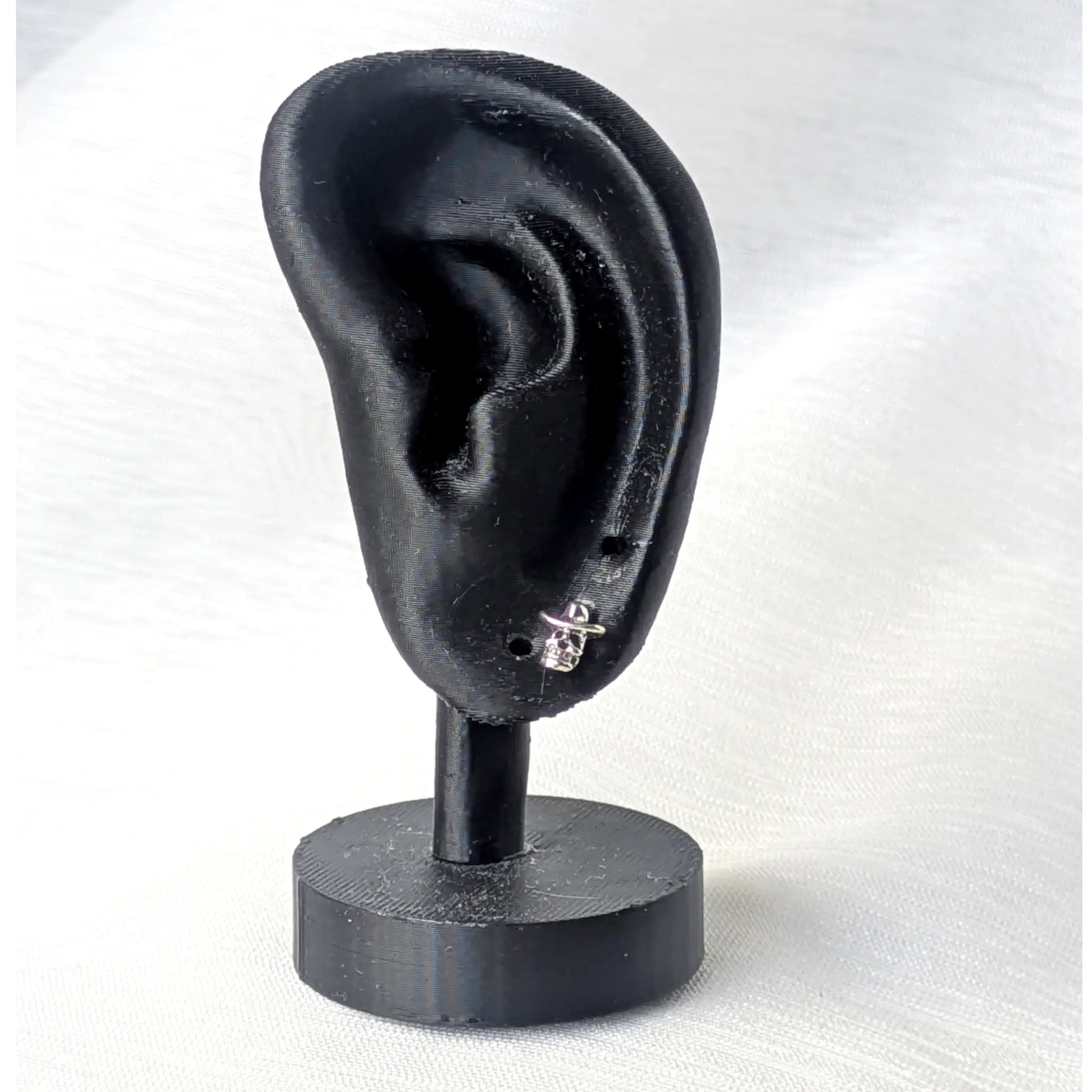 Ein silberner Totenkopf-Ohrstecker mit Cowboyhut getragen an einem mattschwarzen Deko-Ohr zur Größenansicht.