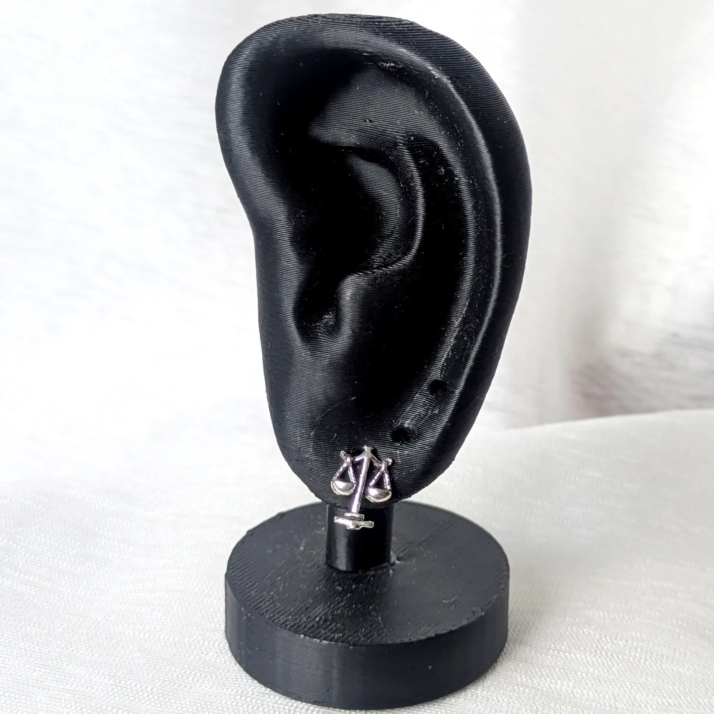 Ein zarter Waage-Ohrstecker aus Silber getragen an einem mattschwarzen Deko-Ohr zur Veranschaulichung der Größe.