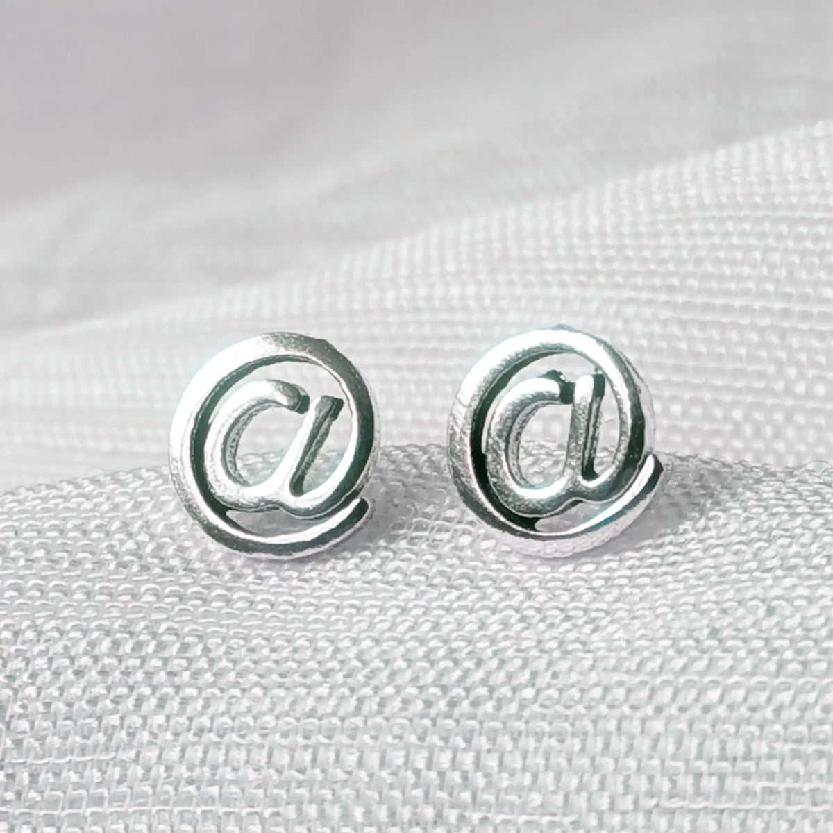 Detailaufnahme glänzender 925 Silber Ohrstecker in Form eines @-Zeichens auf weißem Stoff.
