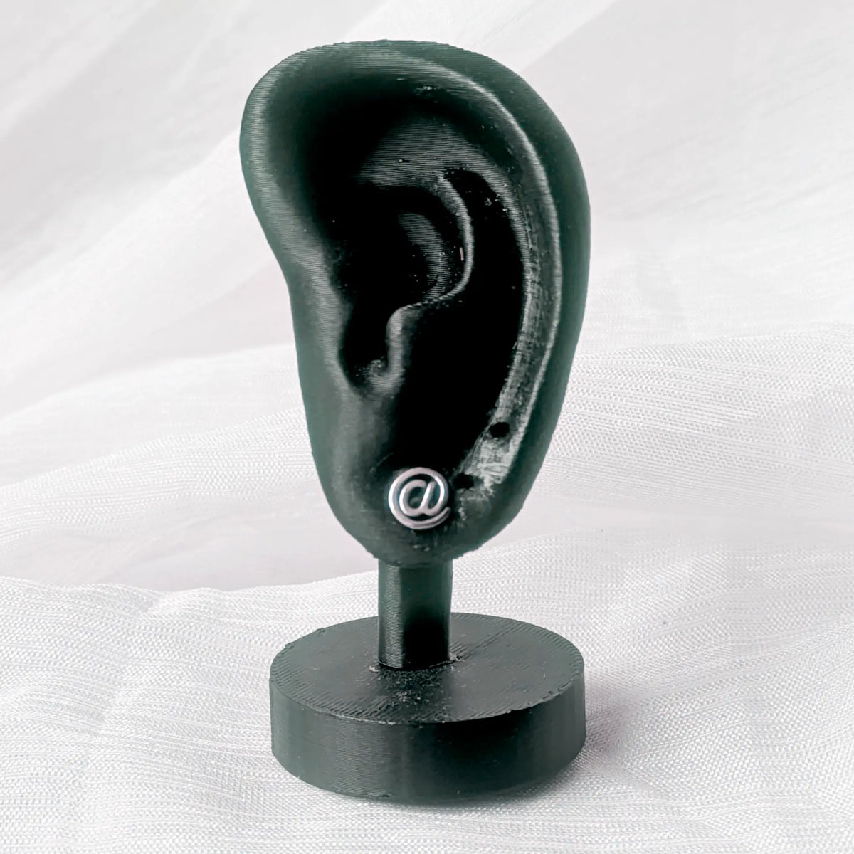 Ein @-Zeichen Silber-Ohrstecker präsentiert an einem schwarzen 3D-Ohr-Display zur Veranschaulichung der Proportionen.